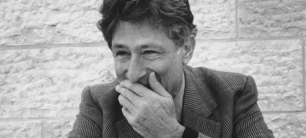 Black and white photograph of Edward W. Said covering his mouth and laughing, in front of a brick wall. 